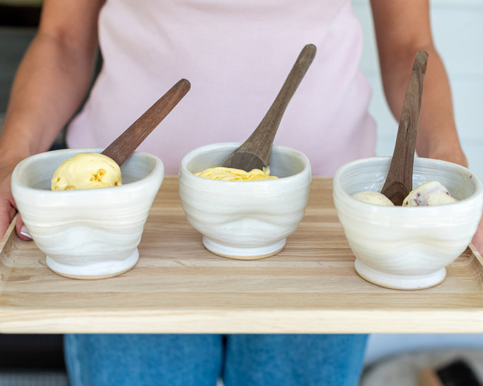 Three small white ceramic bowls with wooden spoons, each containing a scoop of ice cream, arranged on a light wood tray held by a person wearing a pink top and blue jeans.