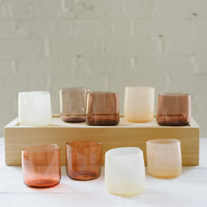 A set of various handcrafted, mouthblown glass cups displayed on a wooden tray against a brick wall background.