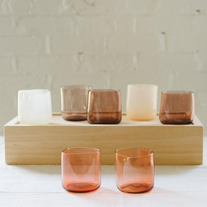 A set of various handcrafted, mouthblown glass cups displayed on a wooden tray against a brick wall background.