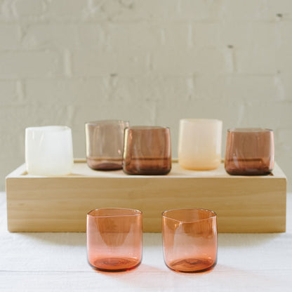 A set of various handcrafted, mouthblown glass cups displayed on a wooden tray against a brick wall background.