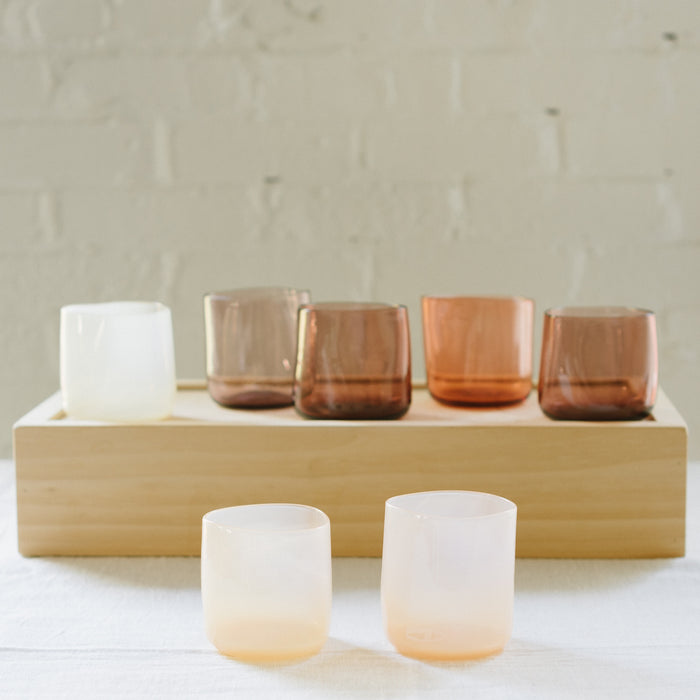 A set of various handcrafted, mouthblown glass cups displayed on a wooden tray against a brick wall background.