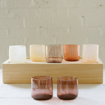 A set of various handcrafted, mouthblown glass cups displayed on a wooden tray against a brick wall background.