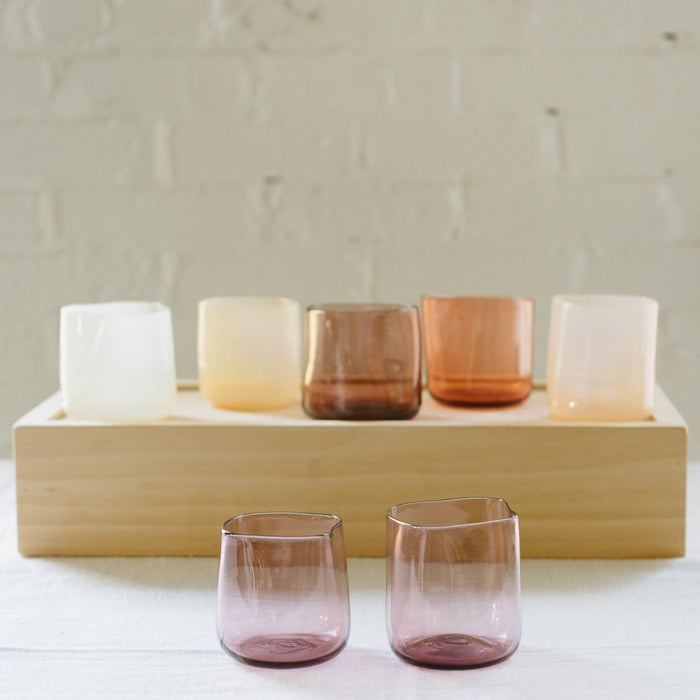 A set of various handcrafted, mouthblown glass cups displayed on a wooden tray against a brick wall background.