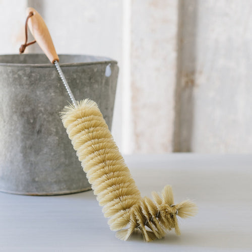 A beige radiator brush with a wooden handle and bristles, resting against a gray metal bucket on a white surface.