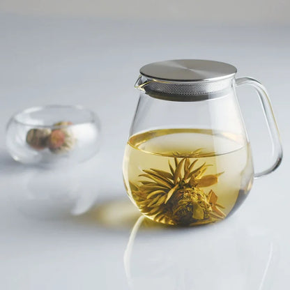 A clear glass teapot with a stainless steel lid and a large curved handle, alongside a small glass cup, on a white background.