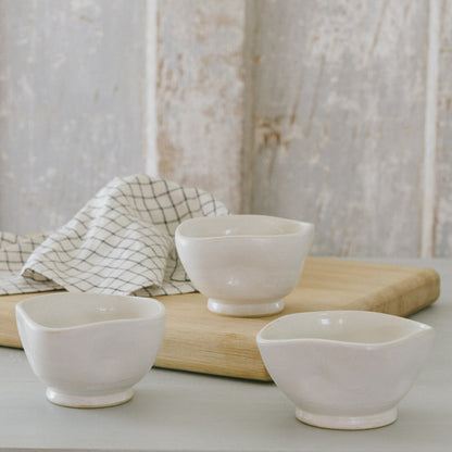 Three white stoneware bowls with white glaze on a wooden surface, with a checkered napkin in the background.